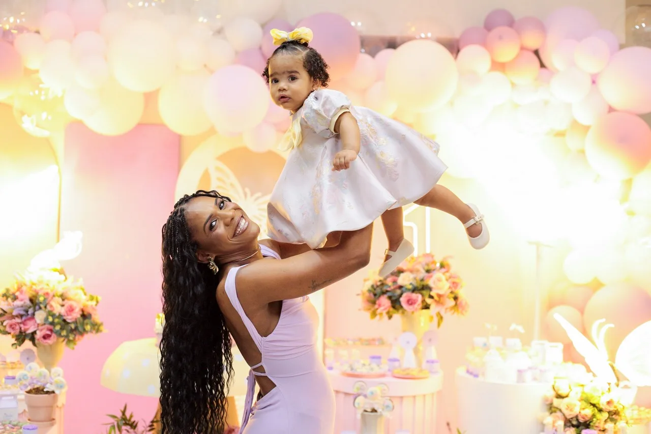 Mulher ergue a menina aniversariante diante da decoração rosa e lilás.