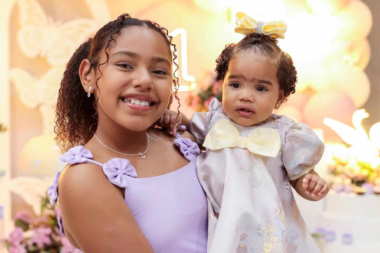Menina sorri com a aniversariante de 1 ano no colo.