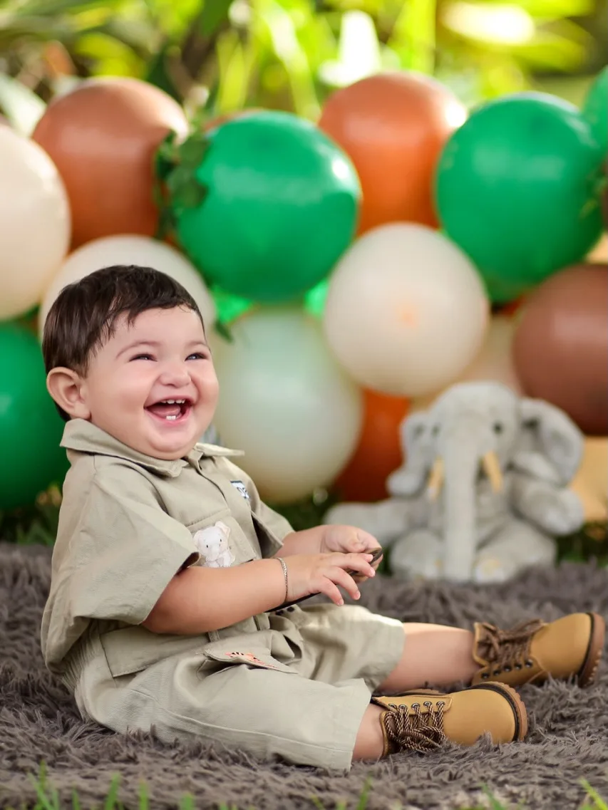 Menino com roupa de safári sorri sentado diante de balões verdes, marrons e creme.