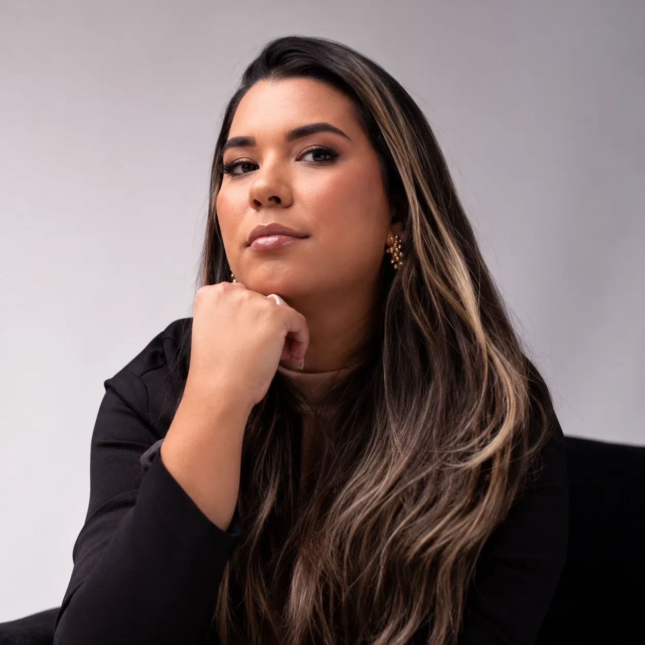 Camila Rodrigues, de cabelos longos com mechas claras e blazer preto, com a mão apoiada no queixo, em fundo cinza-claro.