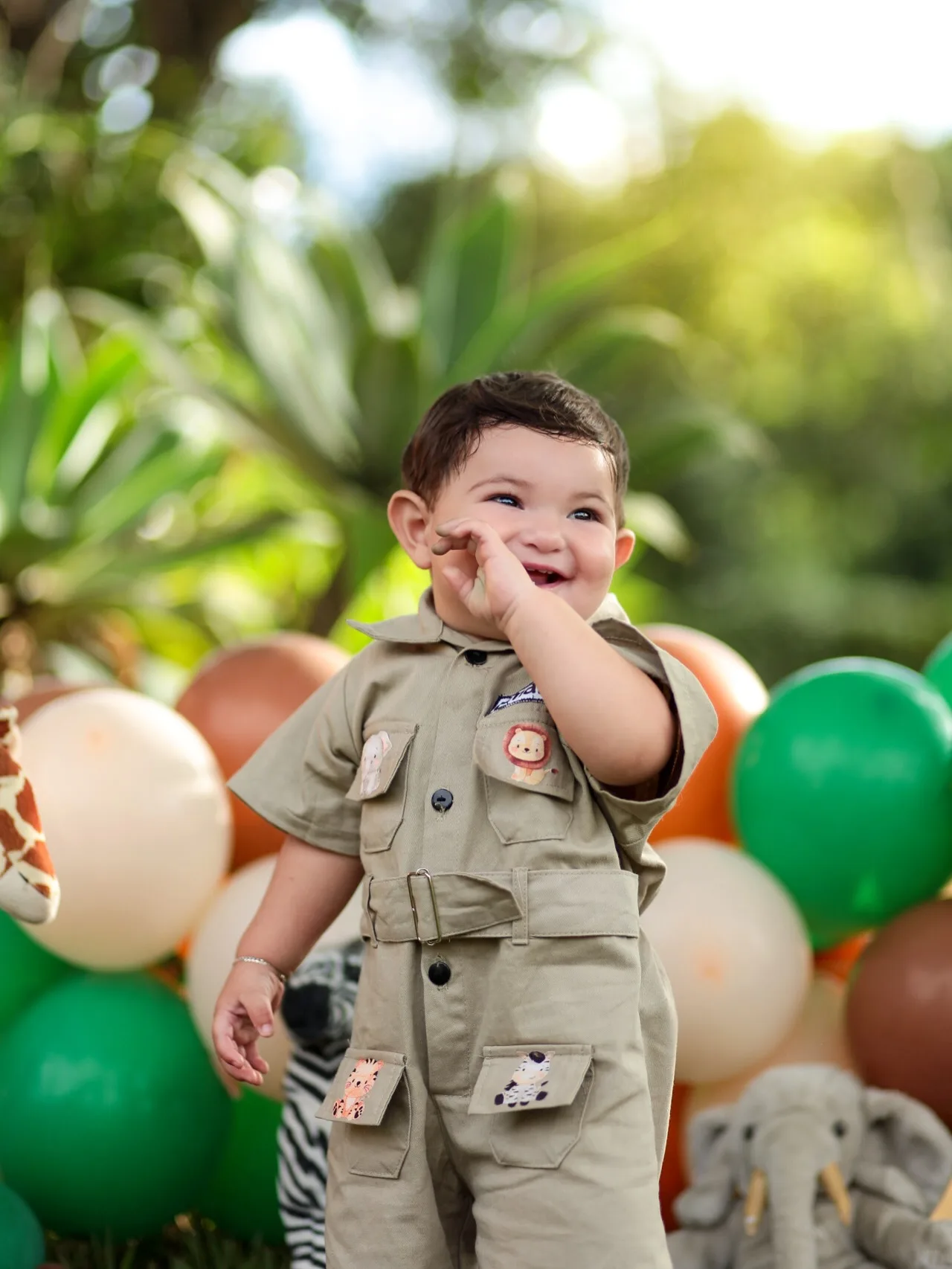 Menino com roupa de safári sorri com a mão no rosto, ao ar livre.