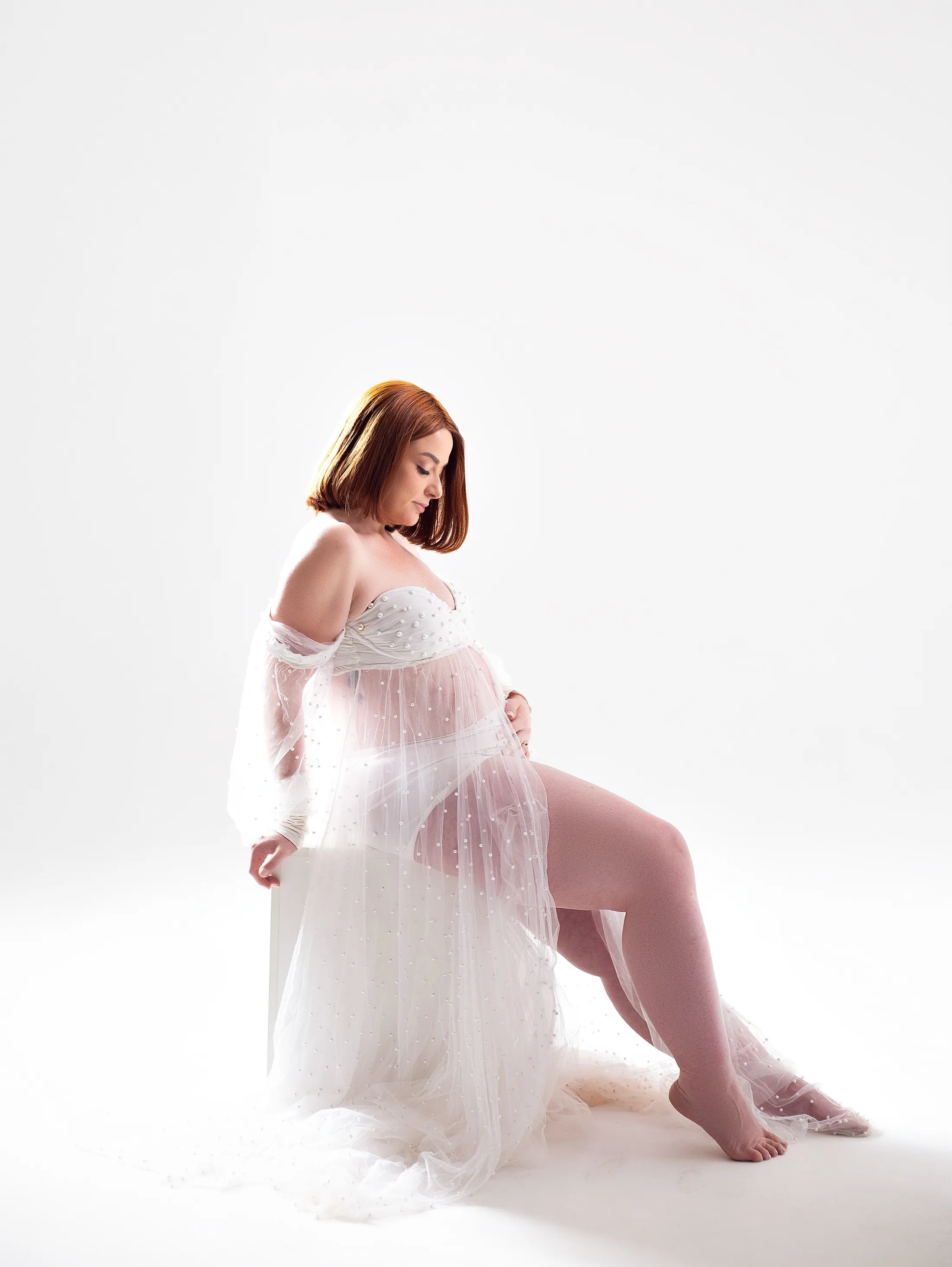 Gestante ruiva de robe branco transparente, sentada, em fundo branco.
