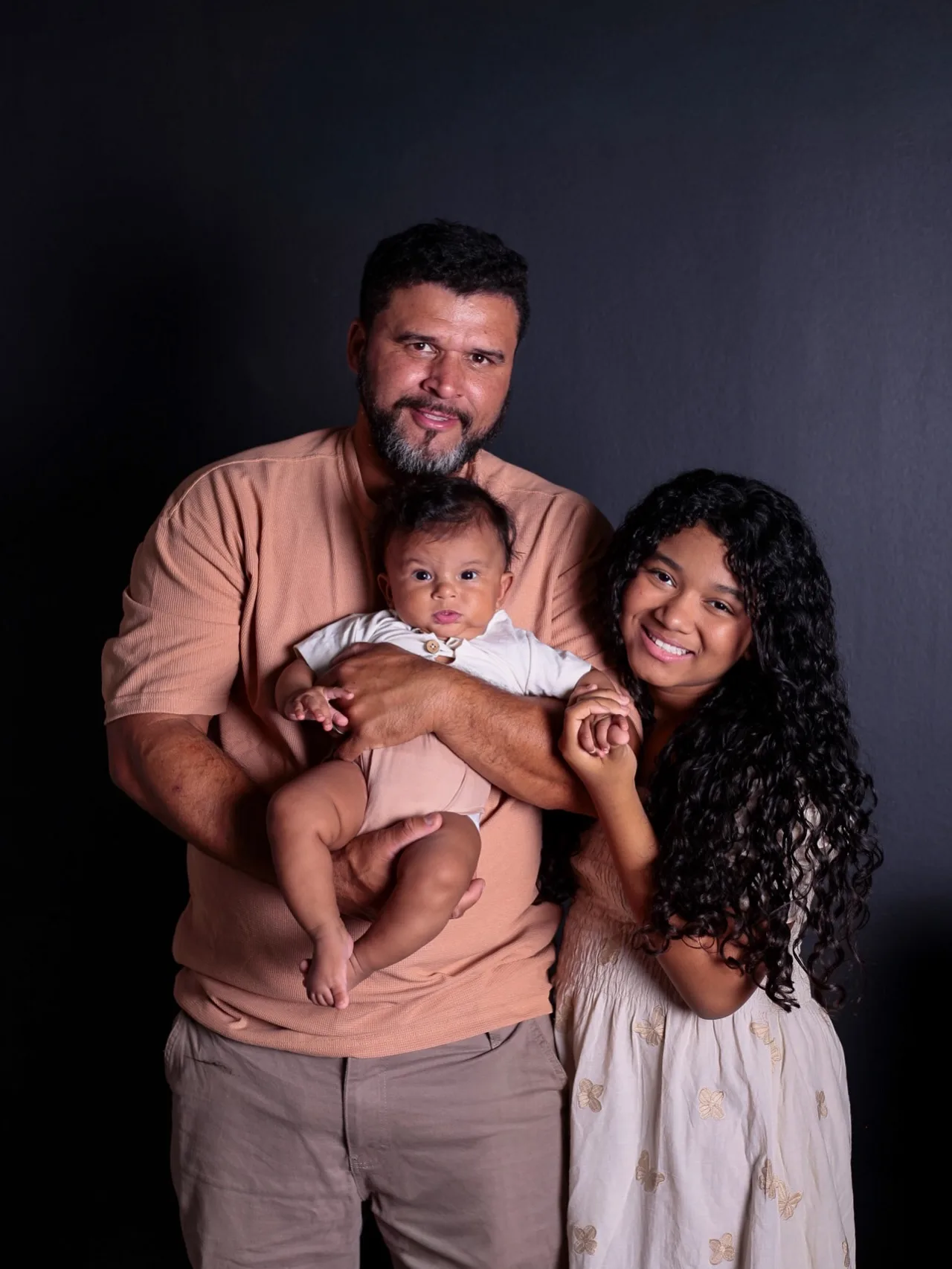 Pai com o bebê no colo e a filha ao lado, diante de um fundo escuro.