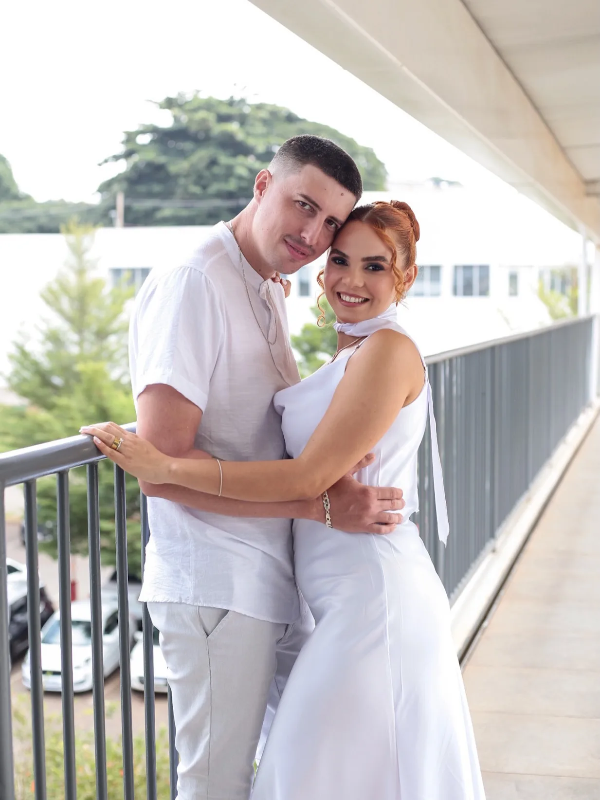 Casal de branco abraçado na varanda, sorrindo para a câmera.
