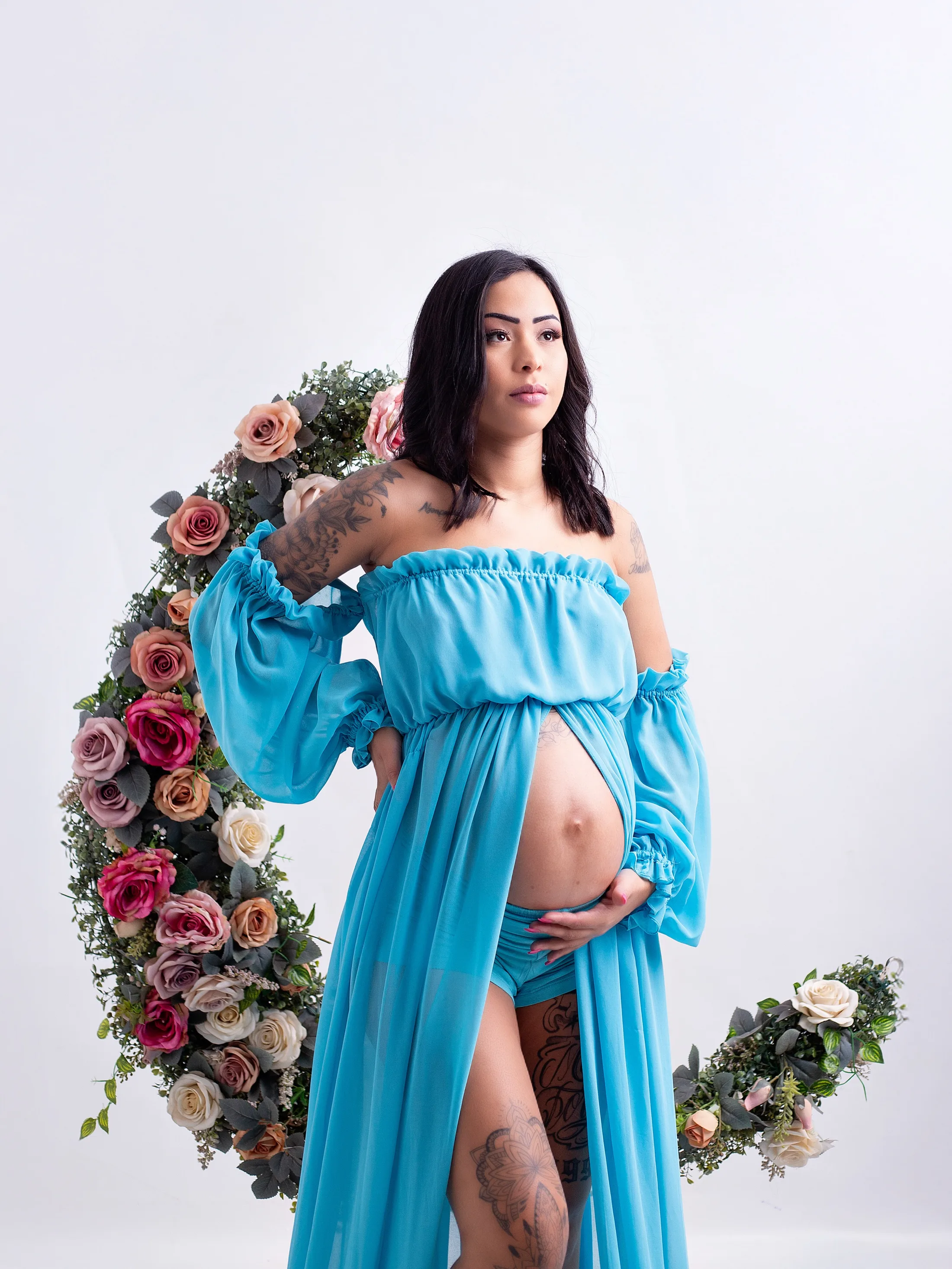 Gestante de vestido azul-turquesa diante de um arco de flores.