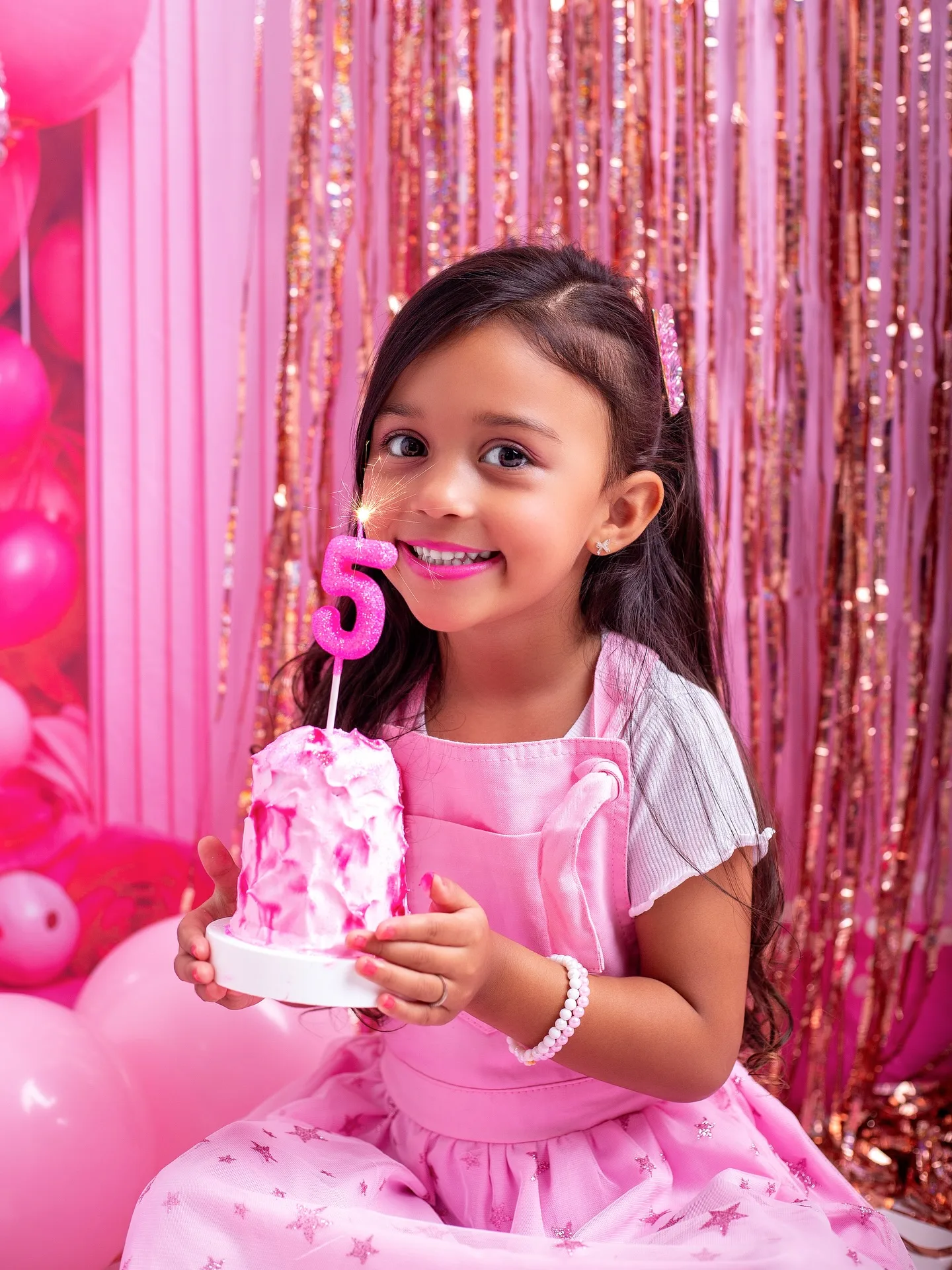Menina segura um bolo com vela de número 5, em cenário rosa.