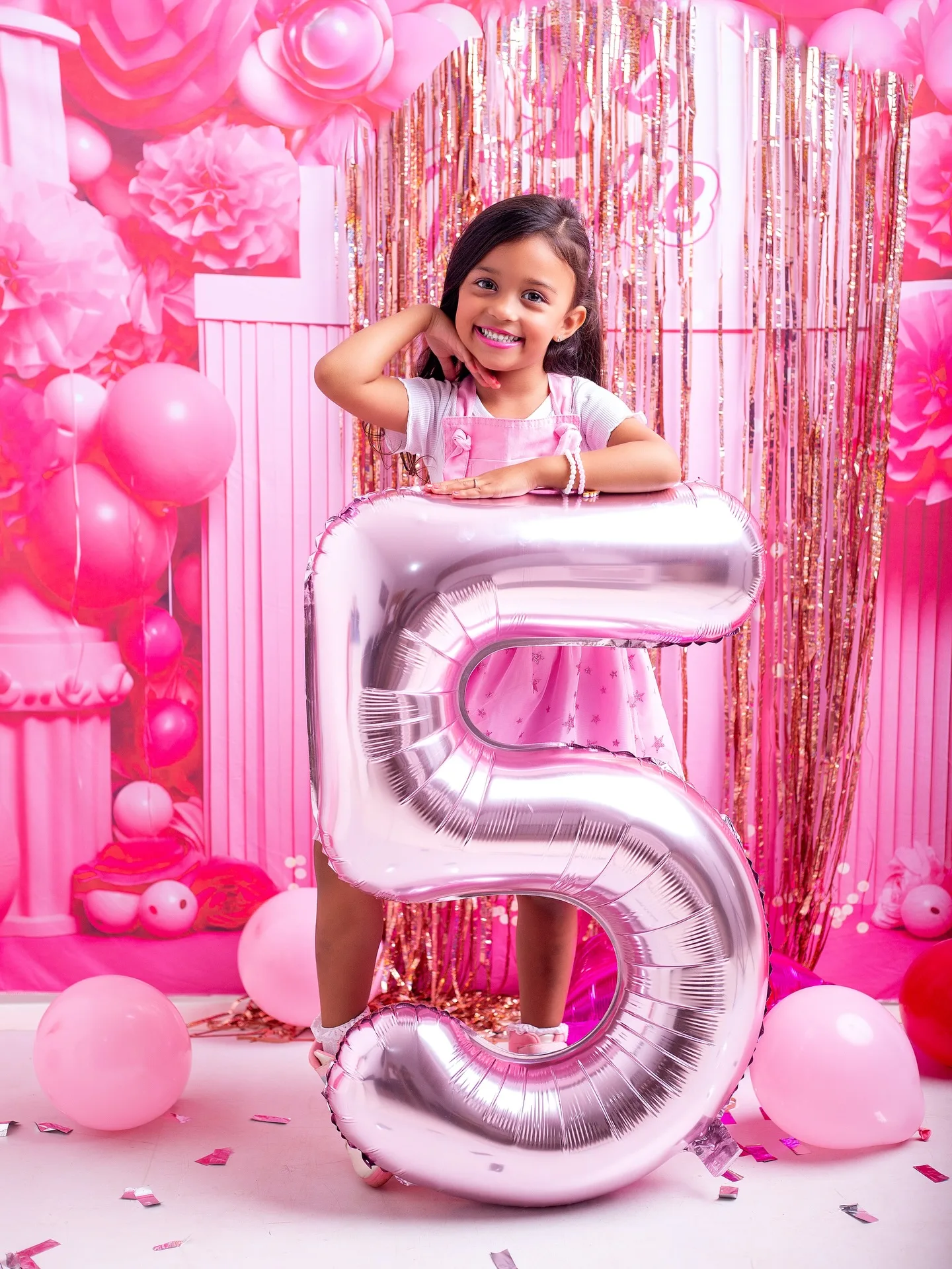 Menina apoiada em um balão prateado de número 5, em cenário rosa.