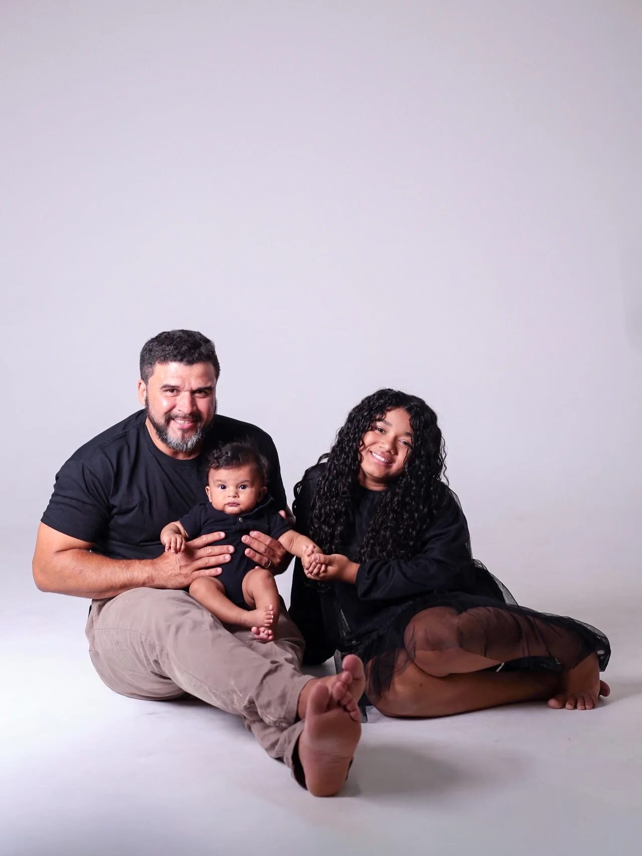 Pai, filha e bebê sentados no chão, em fundo cinza-claro.