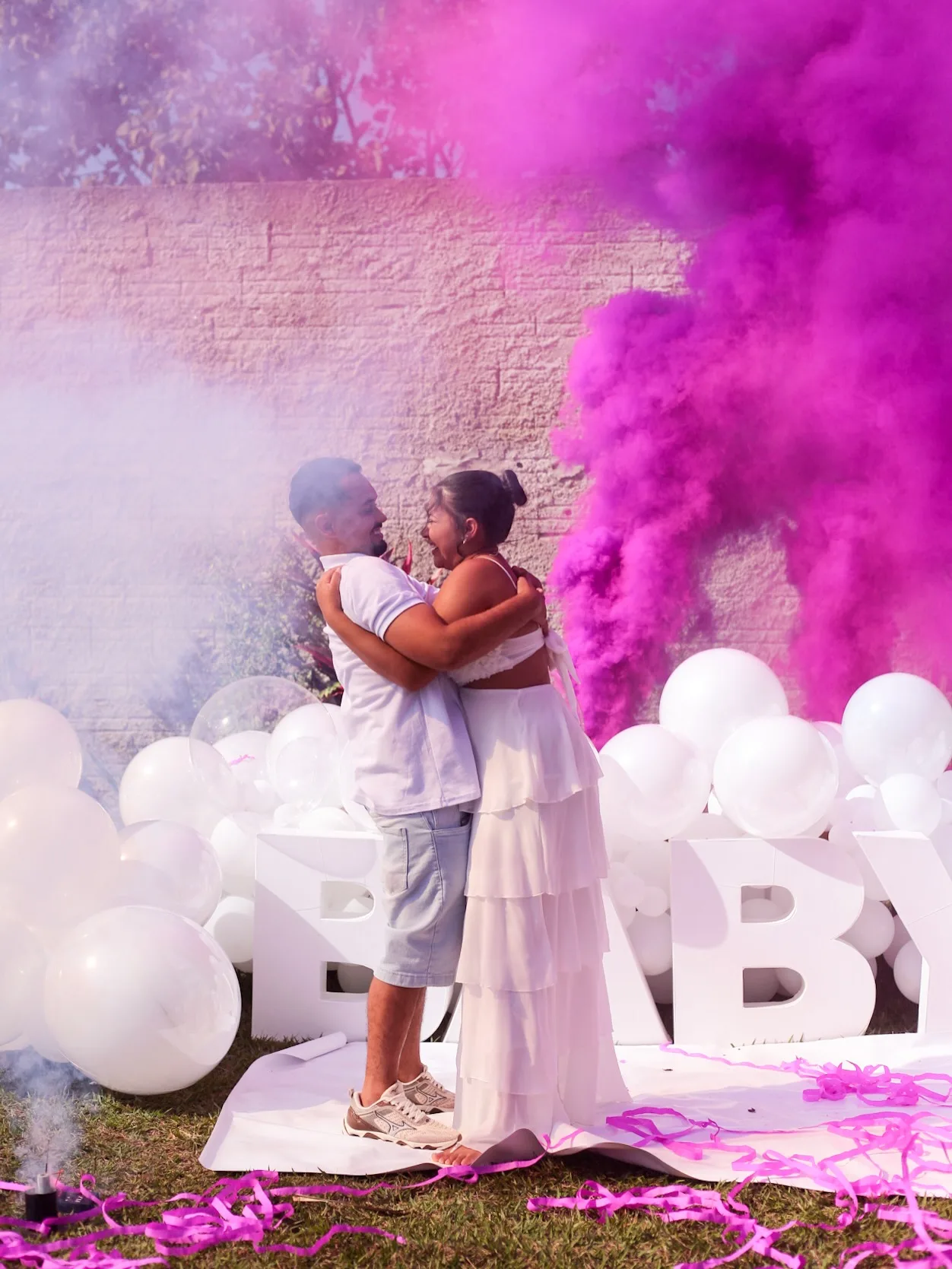 Casal se abraça em meio à fumaça rosa, diante de balões brancos.