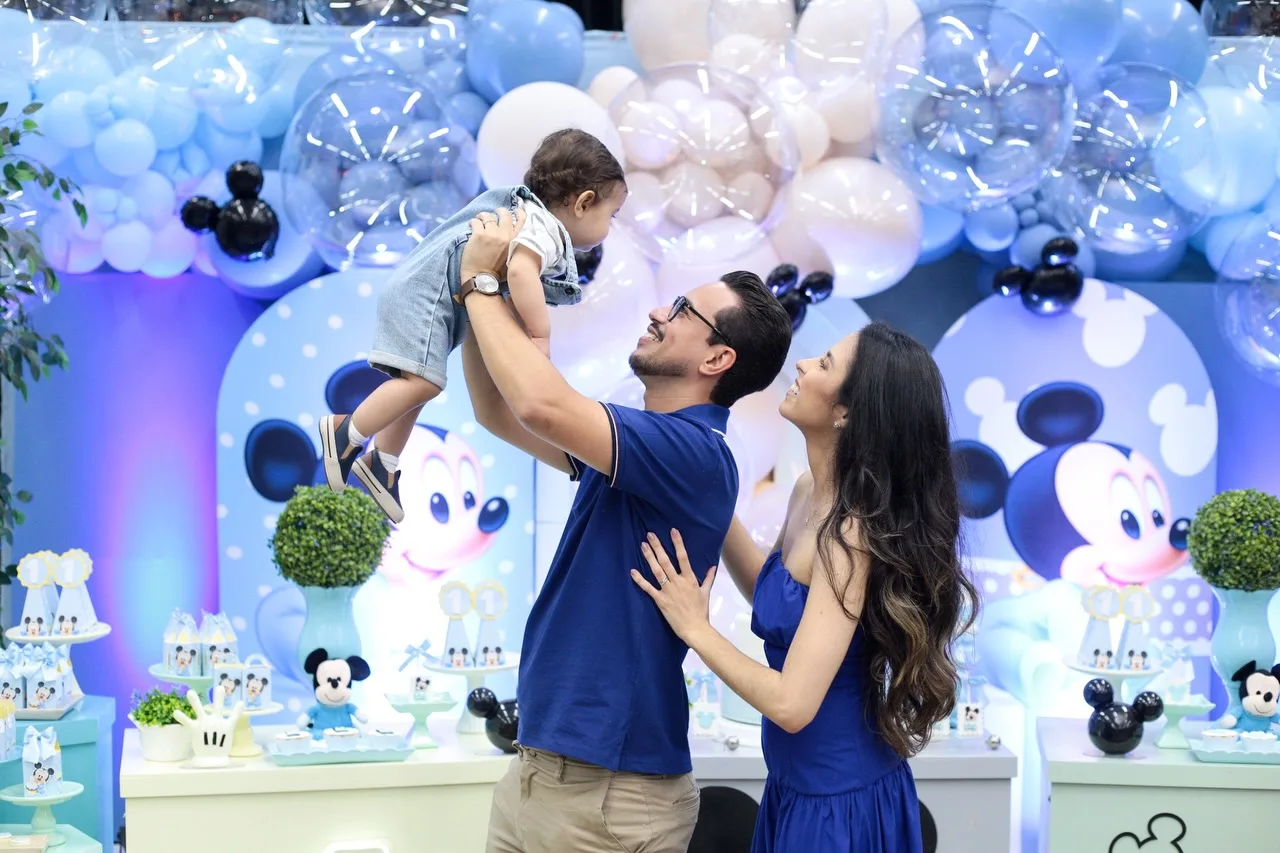 Homem ergue o menino aniversariante diante da decoração azul da festa.