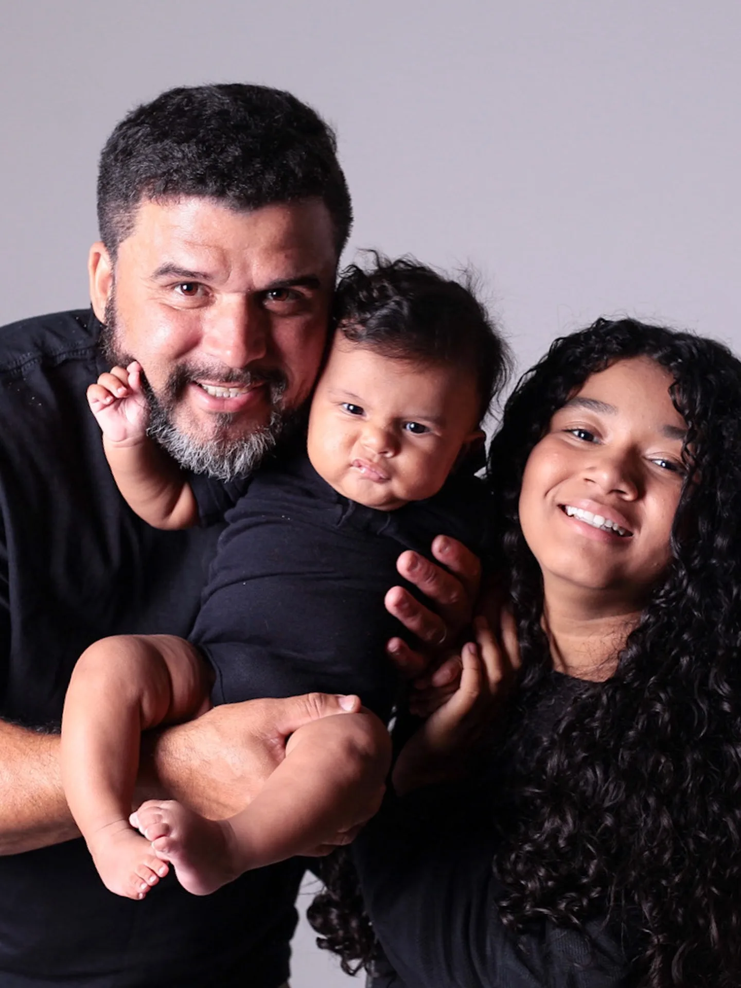 Pai, bebê e filha sorriem em retrato de perto.