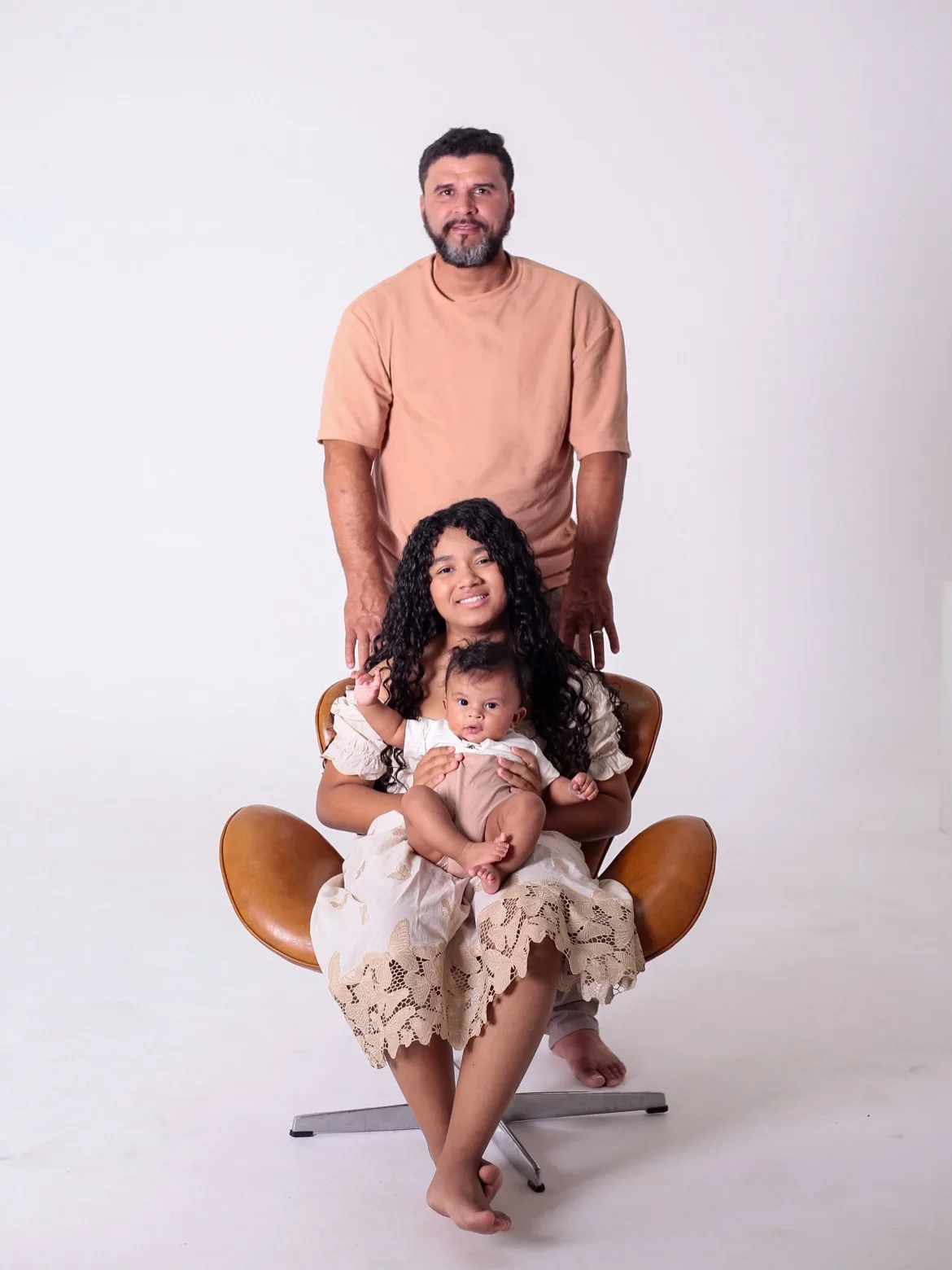 Filha sentada em uma poltrona com o bebê no colo e o pai em pé atrás.