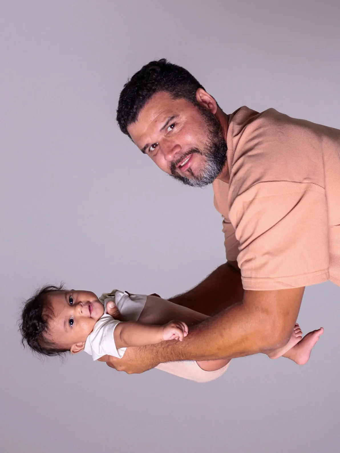 Pai segura o bebê deitado de lado, em fundo cinza-claro.