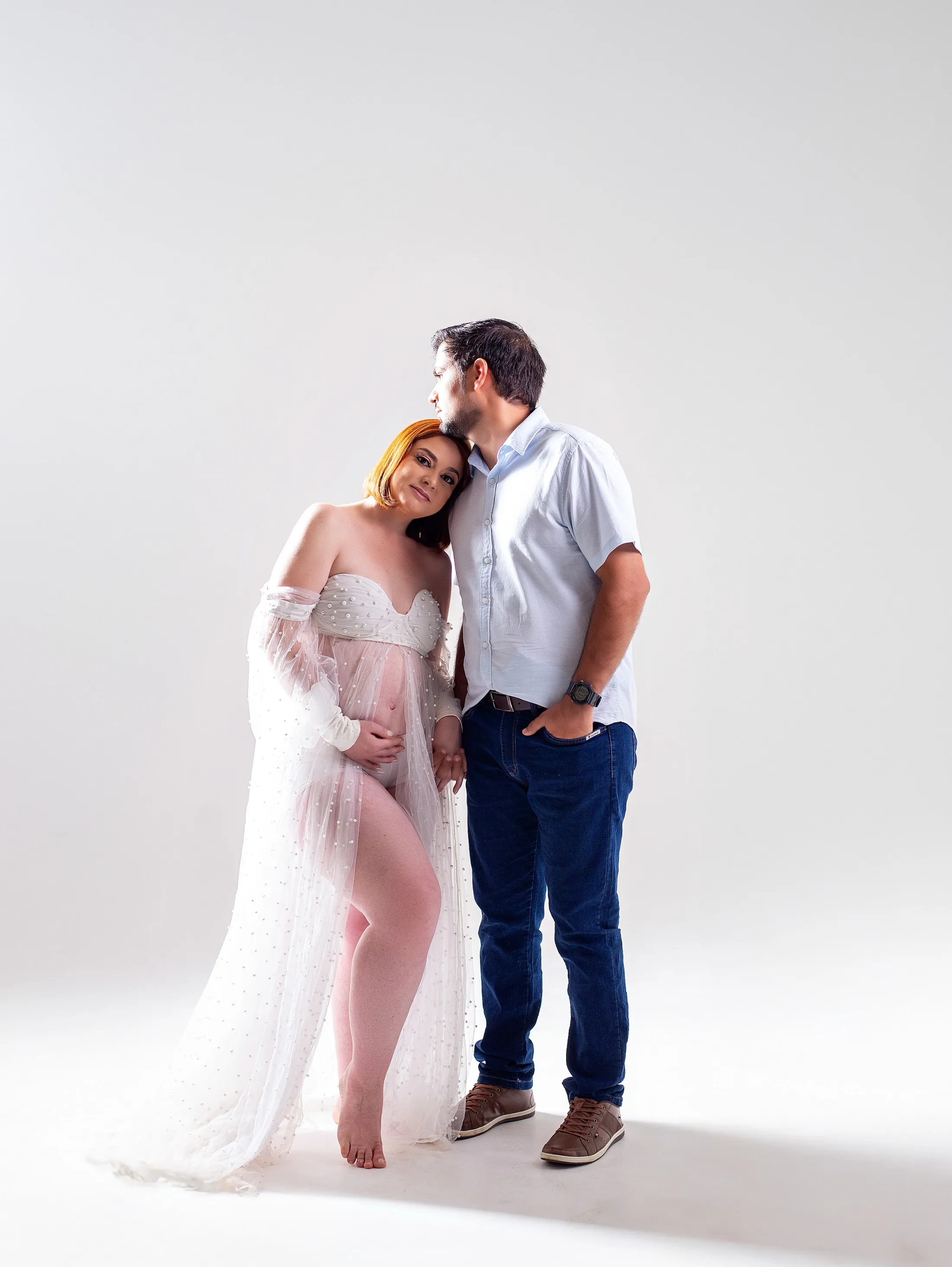 Gestante de robe branco recostada no companheiro de camisa clara, em fundo branco.
