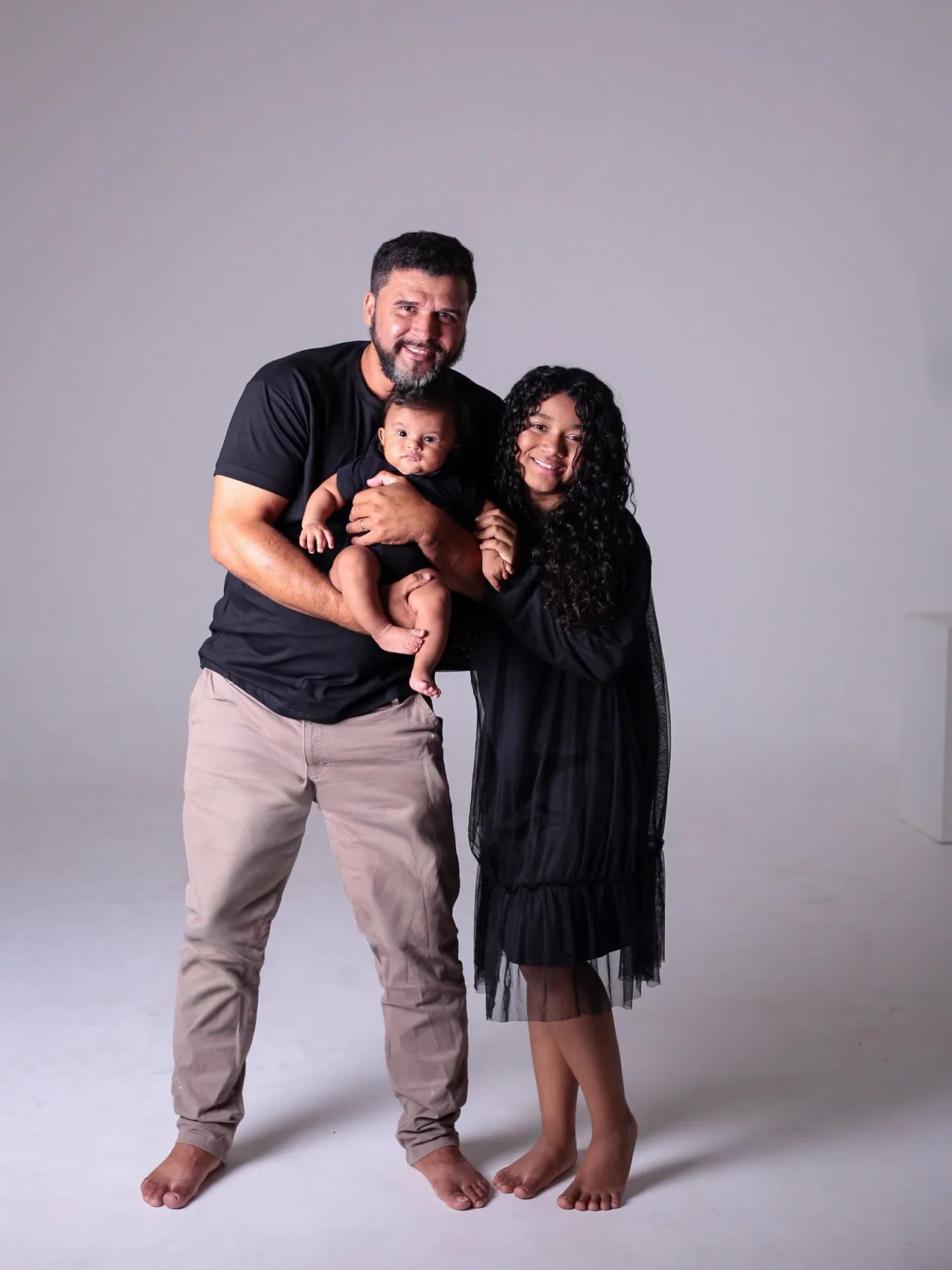 Pai com o bebê no colo ao lado da filha, os três de roupa preta.