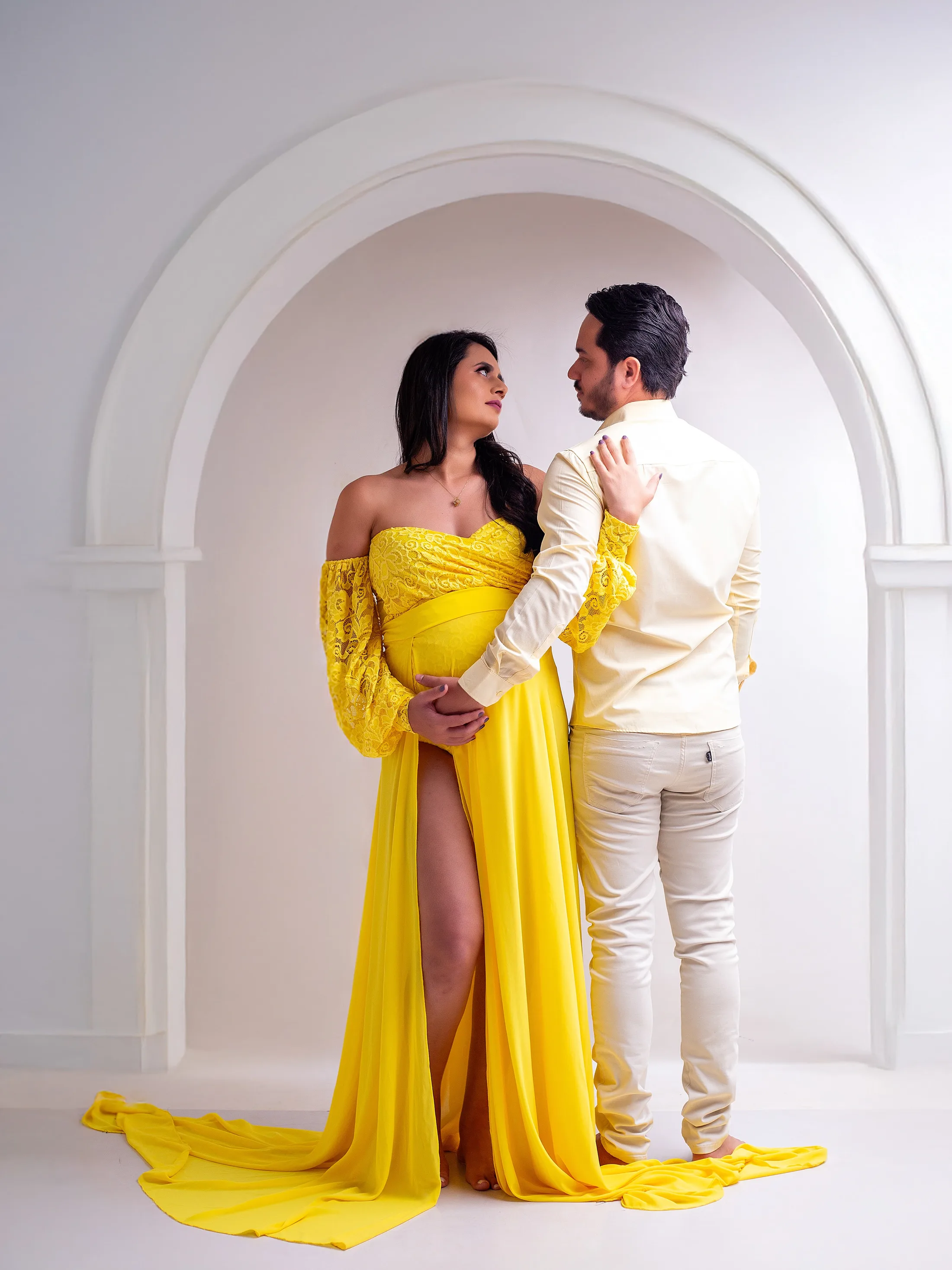 Gestante de vestido amarelo e homem de roupa clara diante de um arco branco.