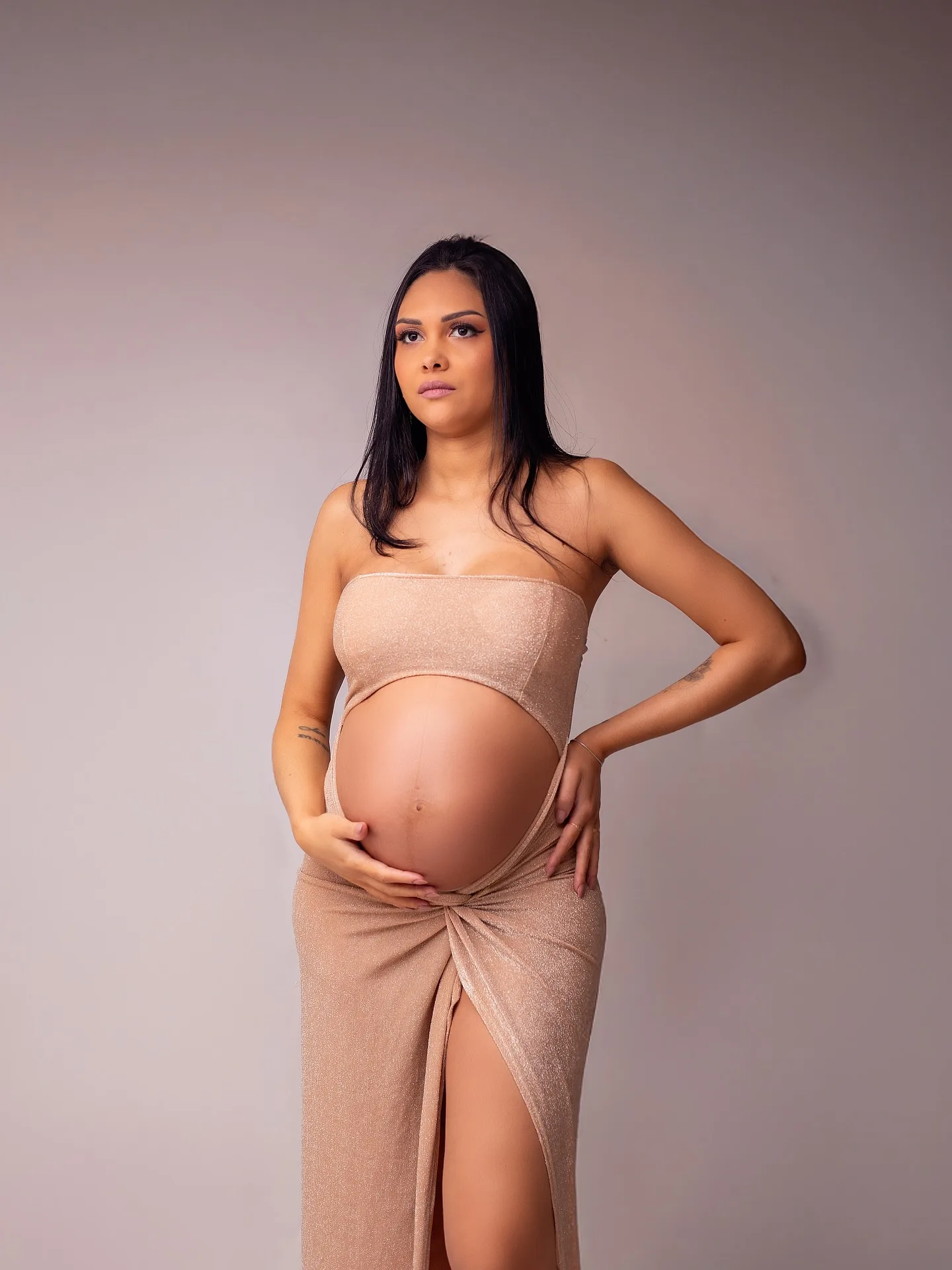 Gestante de top e saia cor de areia com as mãos na barriga, em fundo cinza.