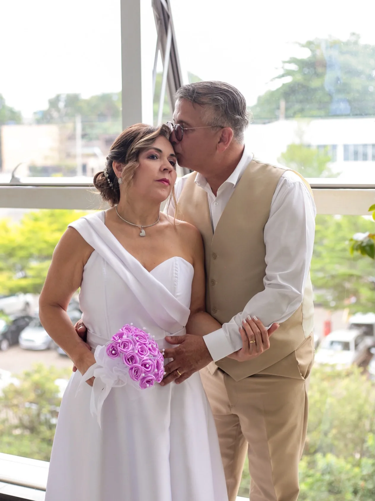 Homem beija a testa da noiva, que segura um buquê lilás, perto de uma janela.