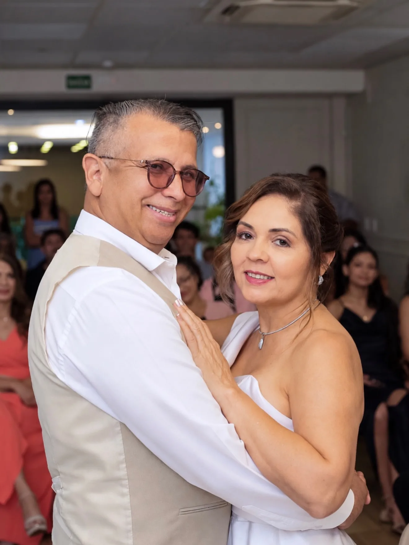 Casal abraçado sorri durante a cerimônia, com convidados ao fundo.