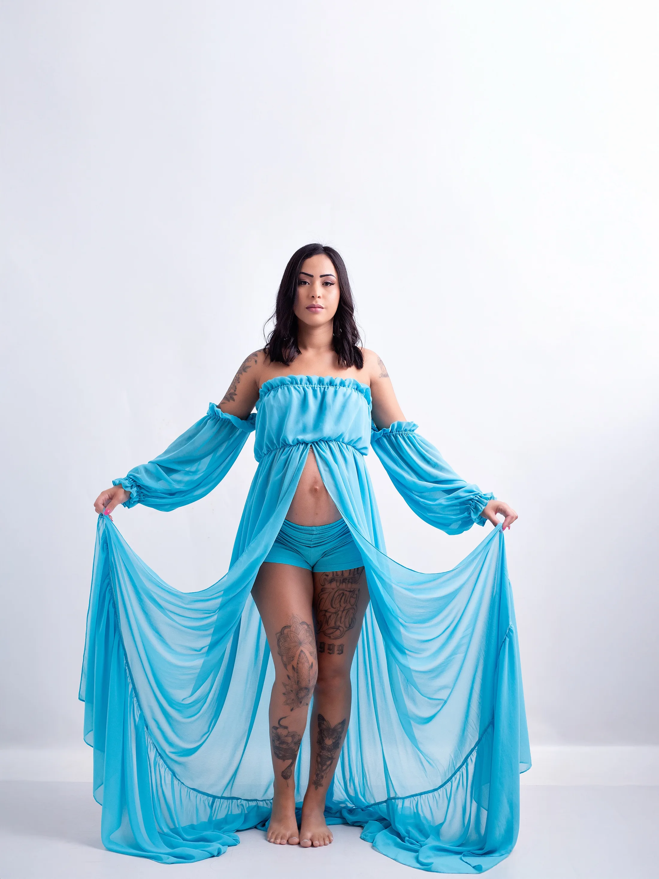 Gestante abre as laterais do vestido azul-turquesa, em fundo branco.