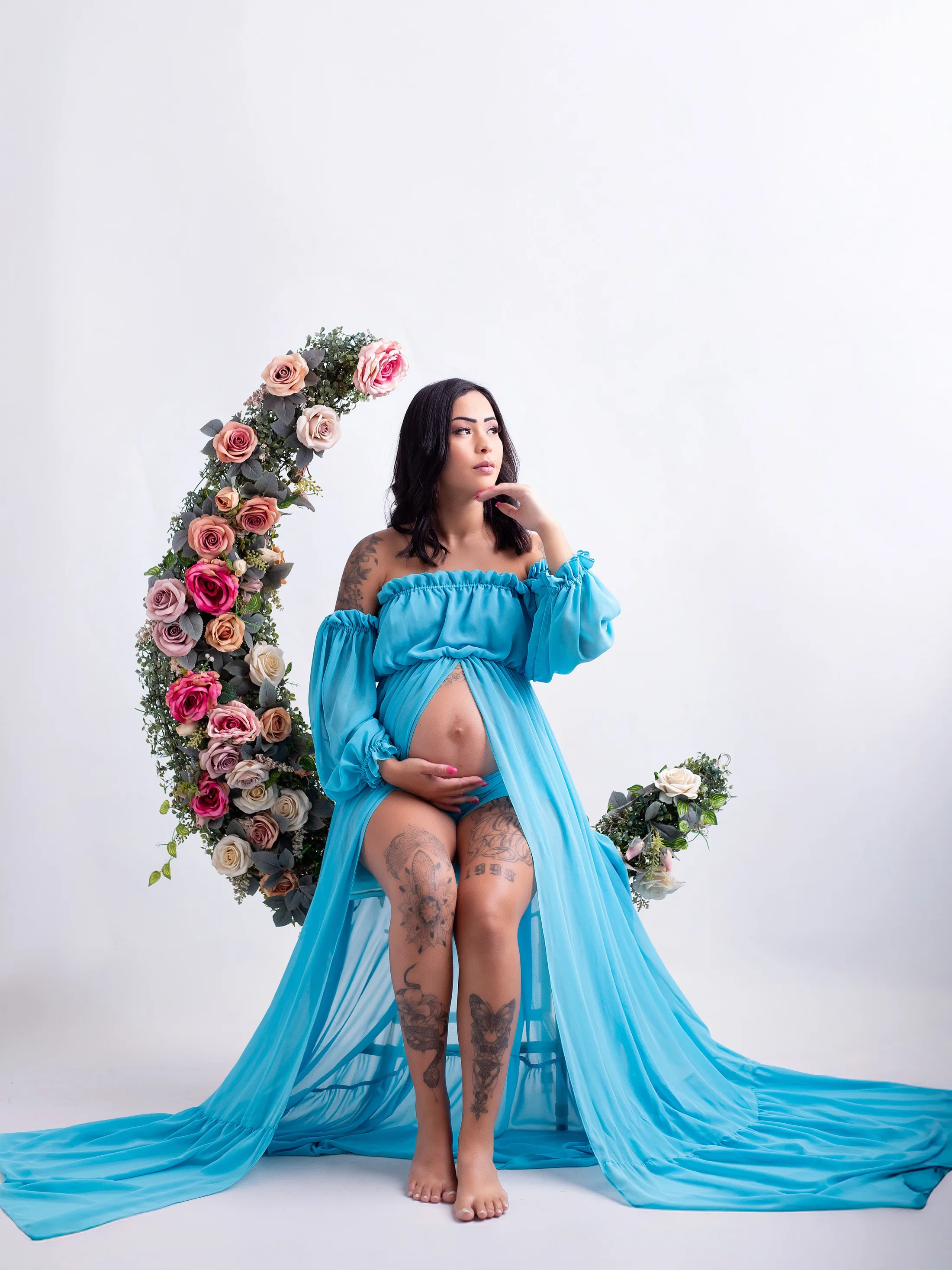 Gestante de vestido azul-turquesa com a mão no queixo, ao lado de um arco de flores.