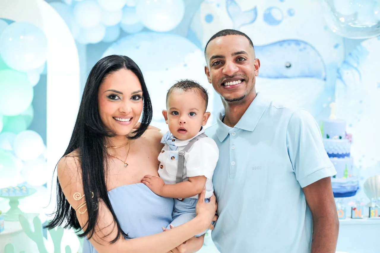 Casal com o menino aniversariante no colo diante da decoração azul-clara.