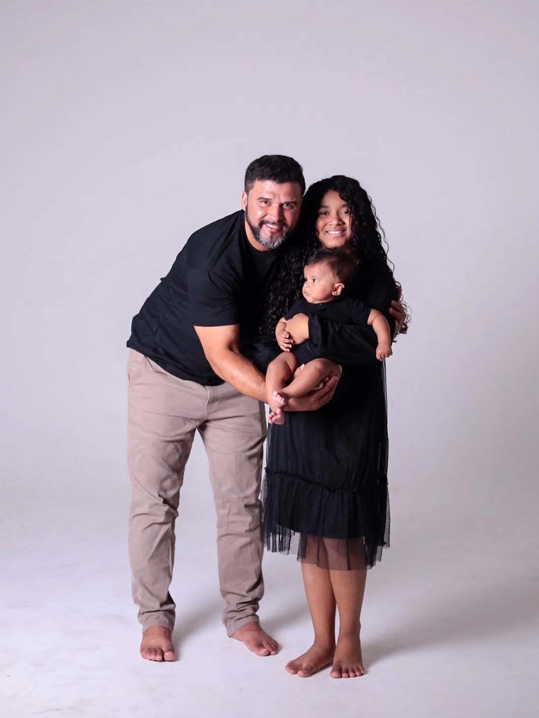 Pai e filha abraçados com o bebê no colo, em fundo cinza-claro.