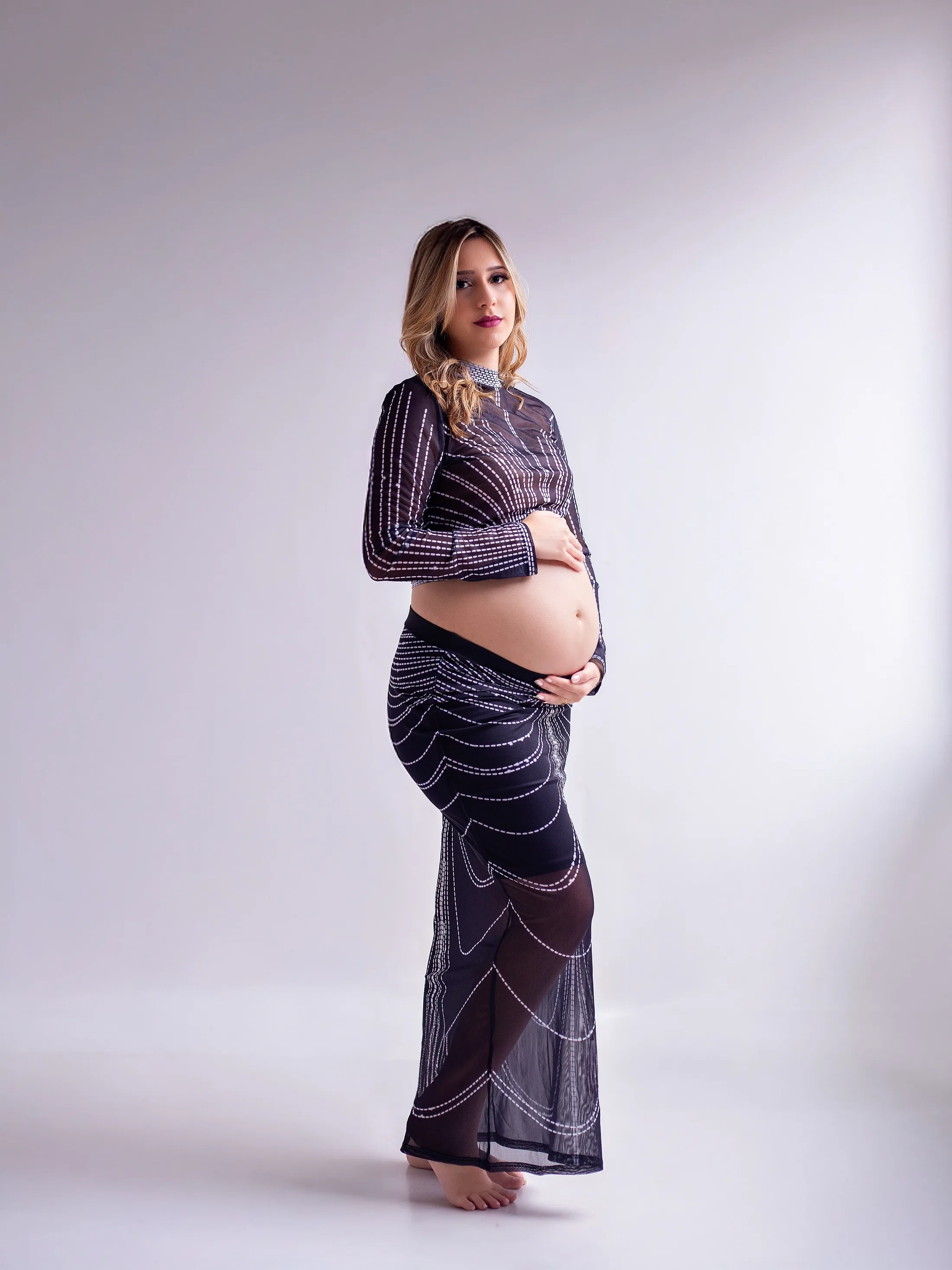 Gestante de conjunto escuro com brilho, de perfil, com a mão na barriga.