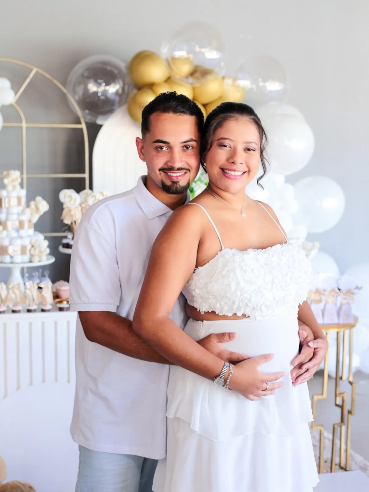 Casal sorri abraçado diante de balões dourados e brancos, no chá revelação.