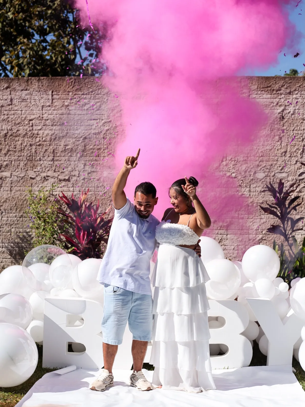 Casal comemora com os braços para cima em meio à fumaça rosa do chá revelação.