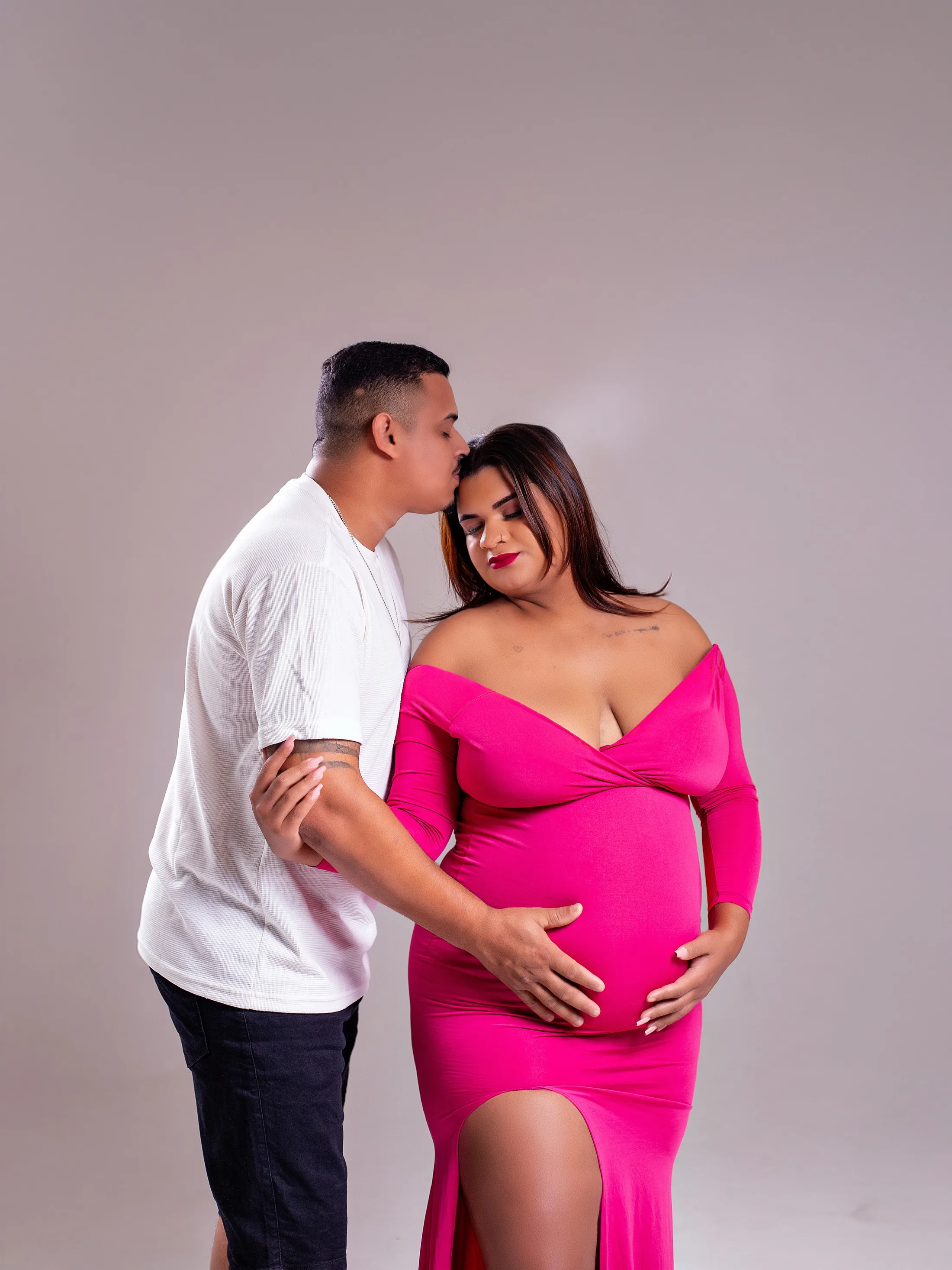 Homem de camiseta branca beija a cabeça da gestante de vestido pink.