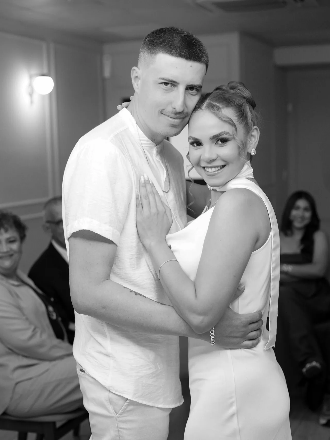 Casal abraçado em foto preto e branco, com convidados ao fundo.