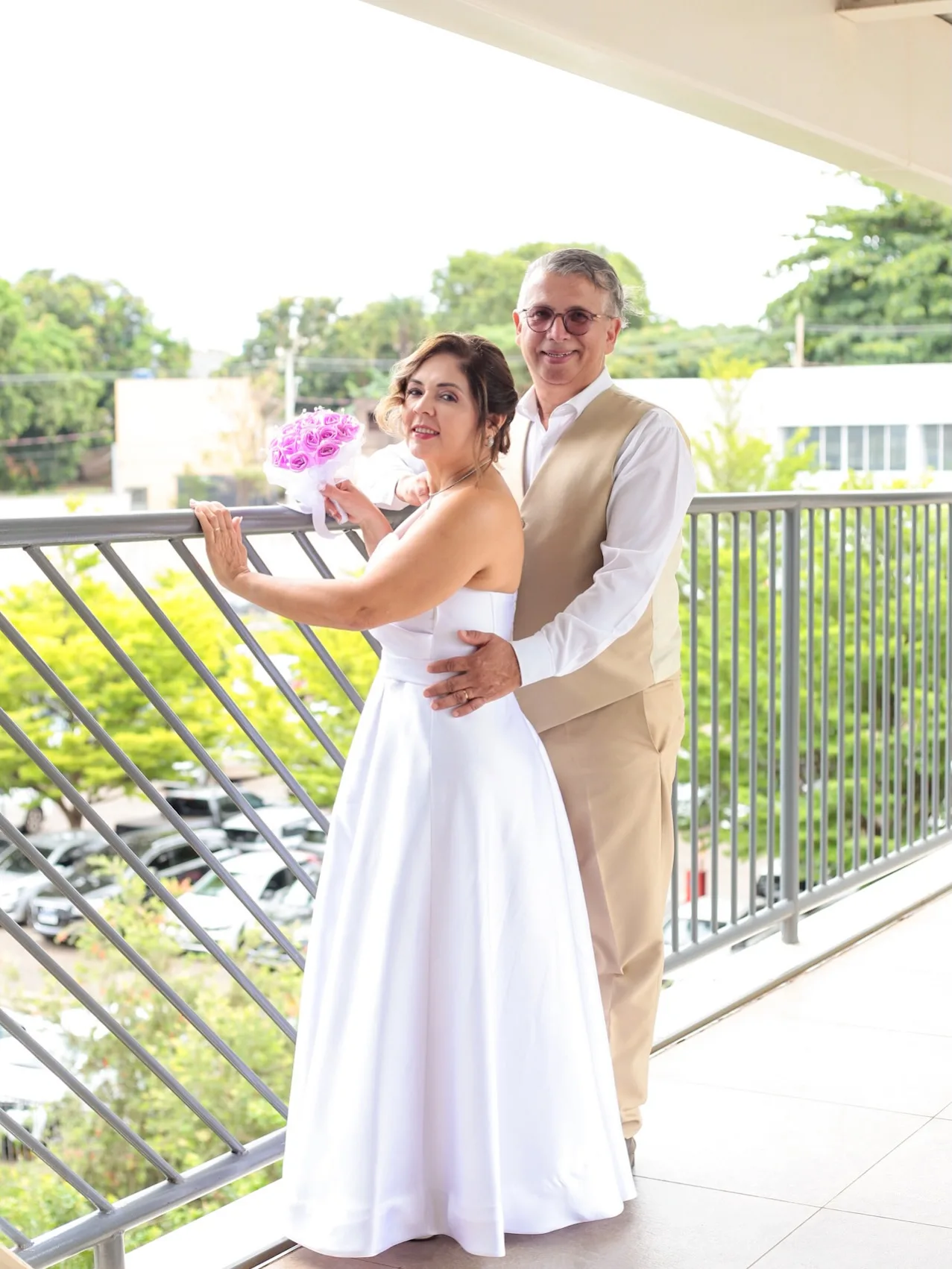 Casal na varanda; a mulher de vestido branco segura um buquê lilás.