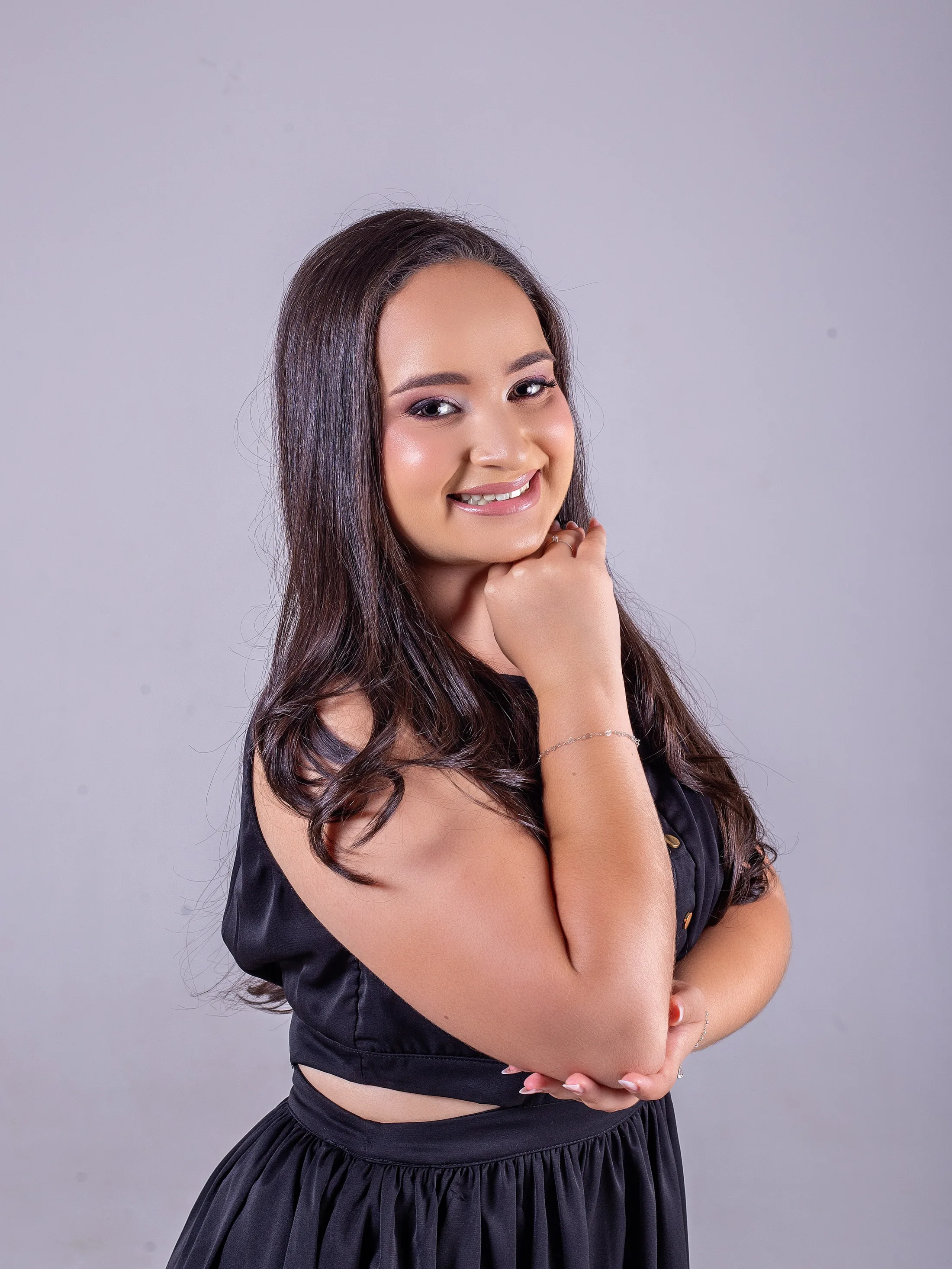Jovem de vestido preto sorri com a mão no queixo, em fundo cinza.